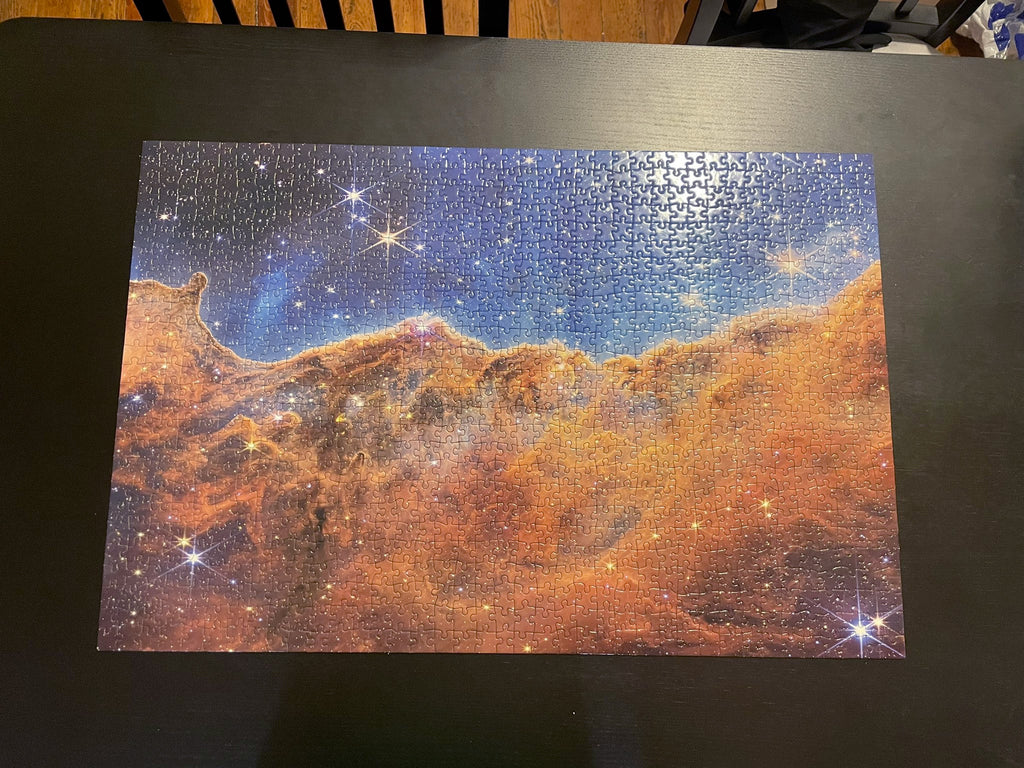 Customer photo by Dr. Luna Zagorac. Image of finished thousand piece puzzle on a black dining table. The theme of the puzzle is the Carina Nebula as images by JWST. The upper third is a blue sky, while the lower two-thirds are a brownish-orange-mauve nebula forming new stars. The entire tableau is littered with stars in the foreground, off different sizes and colors (but predominantly blue or yellow). The larger stars have six spikes coming out of them due to the shape of the telescope’s primary mirror.